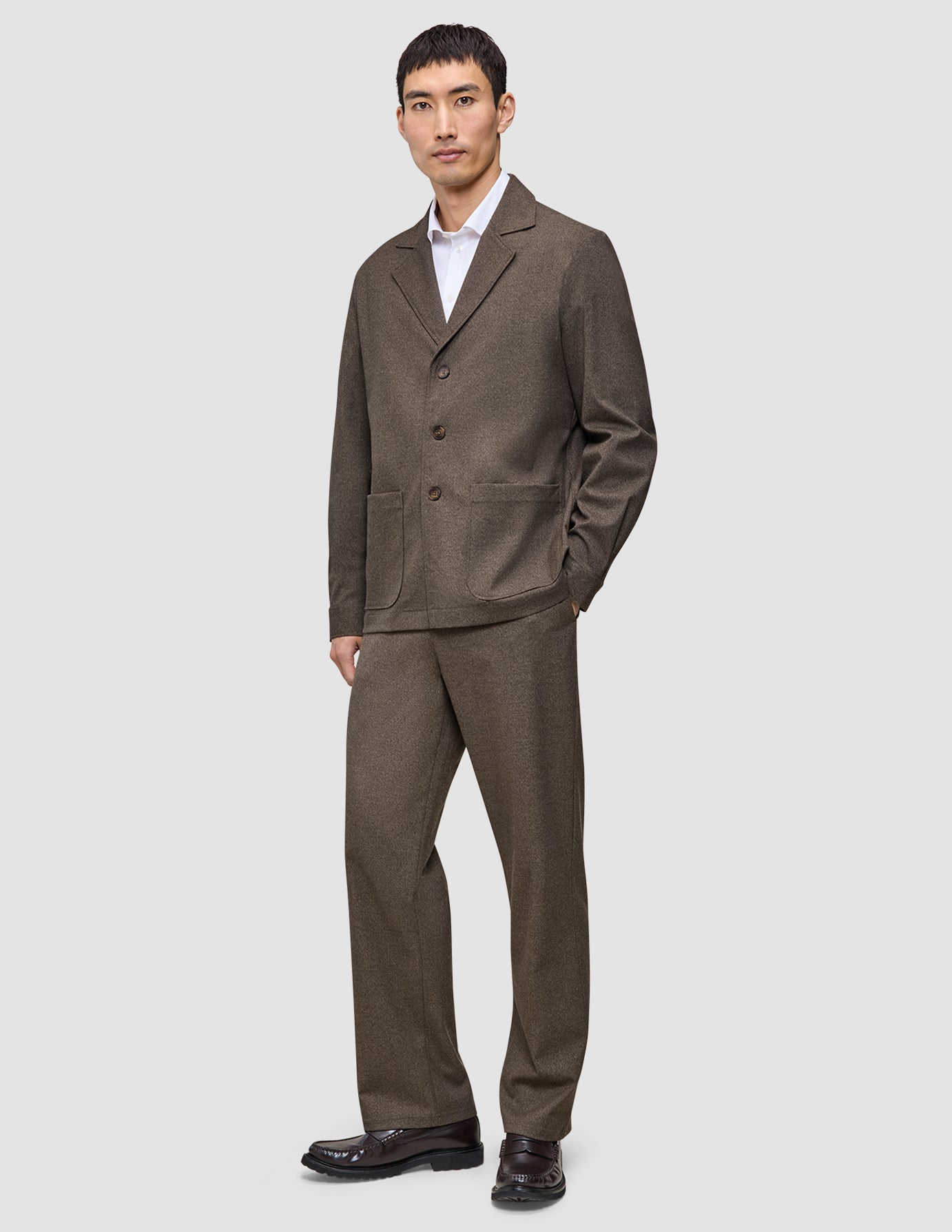 En mand iført en Atelier Overshirt i mørk beige og matchende bukser over en hvid skjorte med krave.